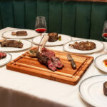 A table full of steaks and wine on a wooden table.