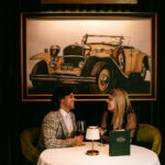 A couple sitting at a table in front of a painting of an old car.